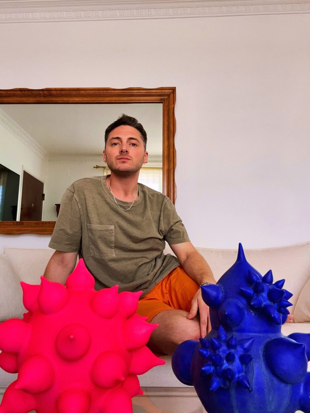 Hugh Remar seated on a white sofa between a hot pink and a cobalt spiked porcelain sculpture